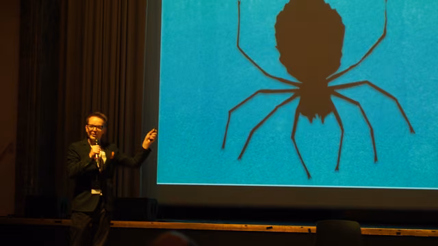 On stage with a mic, pointing at a giant projected spider