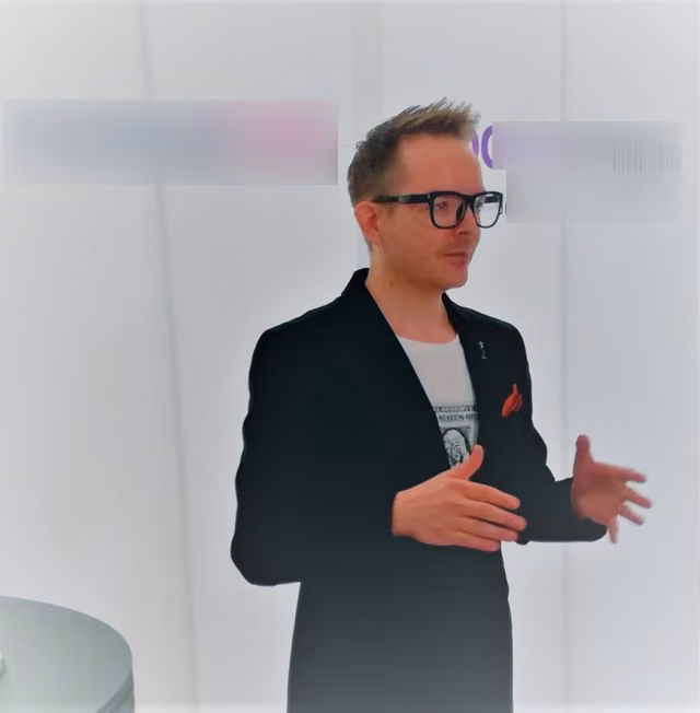 Speaking before an event backdrop in a black blazer with a red pocket square, the backdrop logos blurred