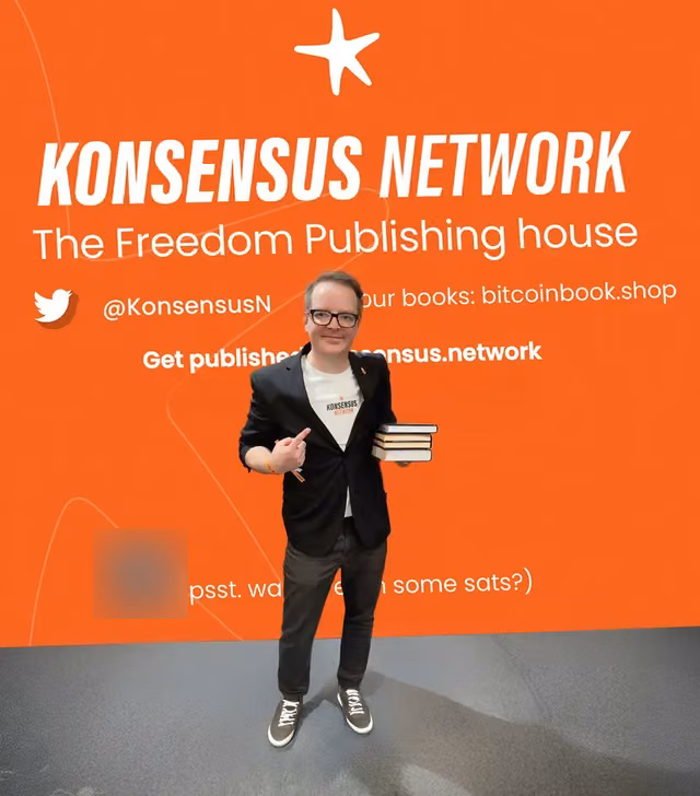 Pointing in front of the orange Konsensus Network backdrop, books in one hand, the QR code blurred