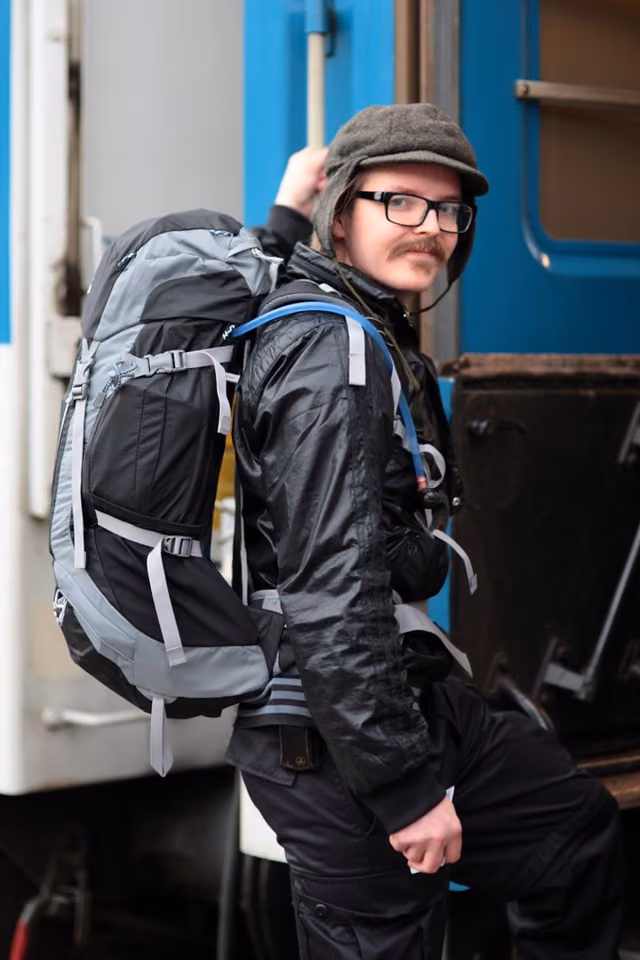 Stepping off a blue-and-white train with a big backpack and a flat cap