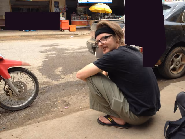 Squatting on a dusty street in Cambodia, in a headband, glasses and flip-flops, the bystanders printed as silhouettes