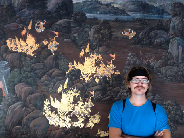 Arms crossed before a gold-leaf temple mural, in a blue tee and a headband