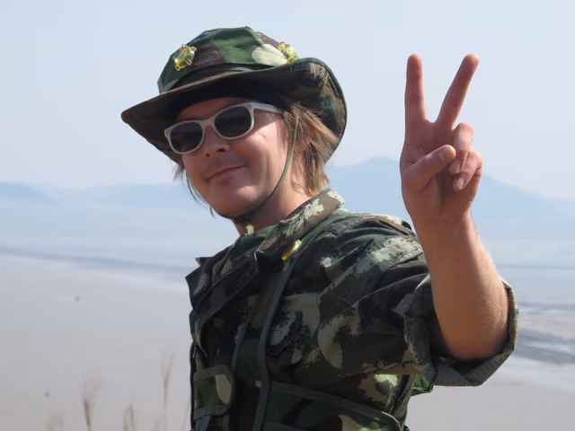 A peace sign against a pale sky, in a camo jacket, a boonie hat and white sunglasses